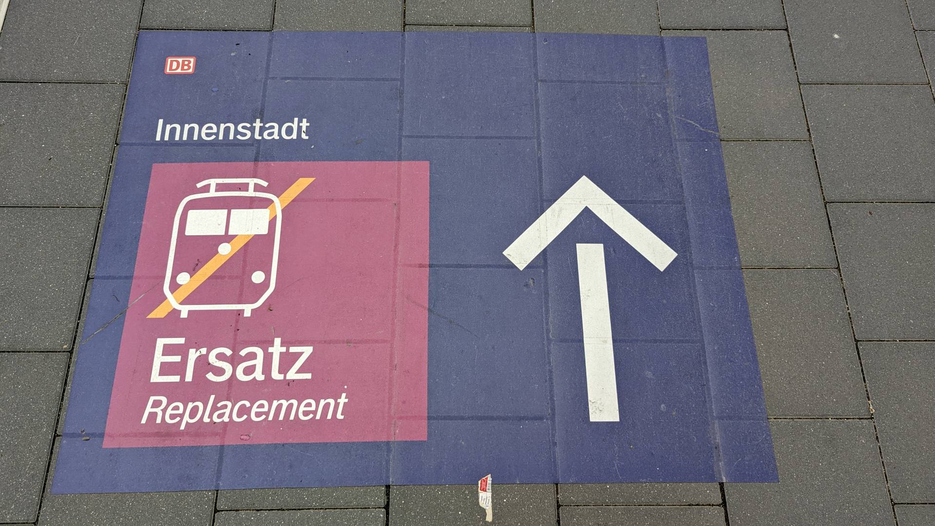 Ein blaues Schild mit einem Symbol für einen Zug, dass durchgestrichen ist. Darüber steht "Innenstadt" und rechts daneben ein großer Pfeil nach vorne als Richtunsgangabe. Oben links in der Ecke ist das Logo von der deutschen Bahn.