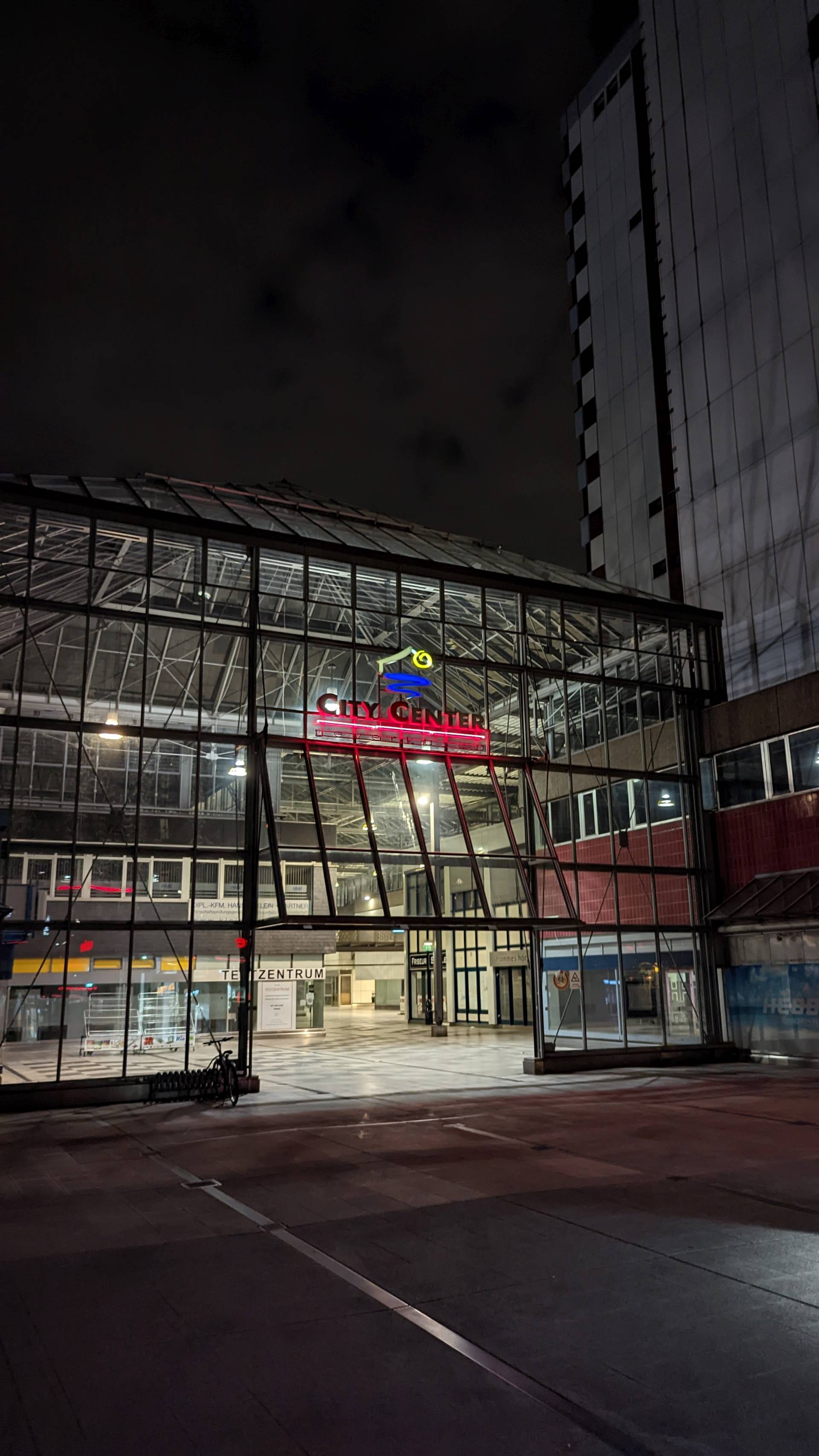 Das City Center in Leverkusen-Mitte bei Nacht. Es ist dunkel, das City Center selbst ist hell beleuchtet. Über dem Eingang zum überdachten Fußgängerbereich leuchtet der Schriftzug "City Center" samt dem Logo