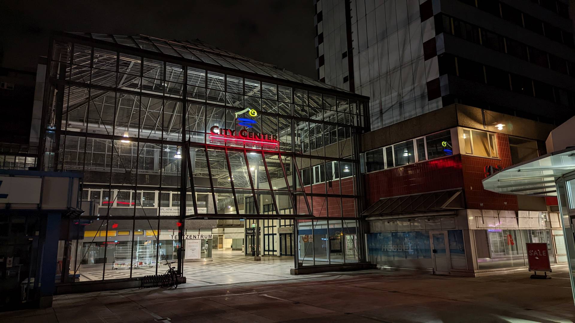 Das City Center in Leverkusen-Mitte bei Nacht. Es ist dunkel, das City Center selbst ist hell beleuchtet. Über dem Eingang zum überdachten Fußgängerbereich leuchtet der Schriftzug "City Center" samt dem Logo