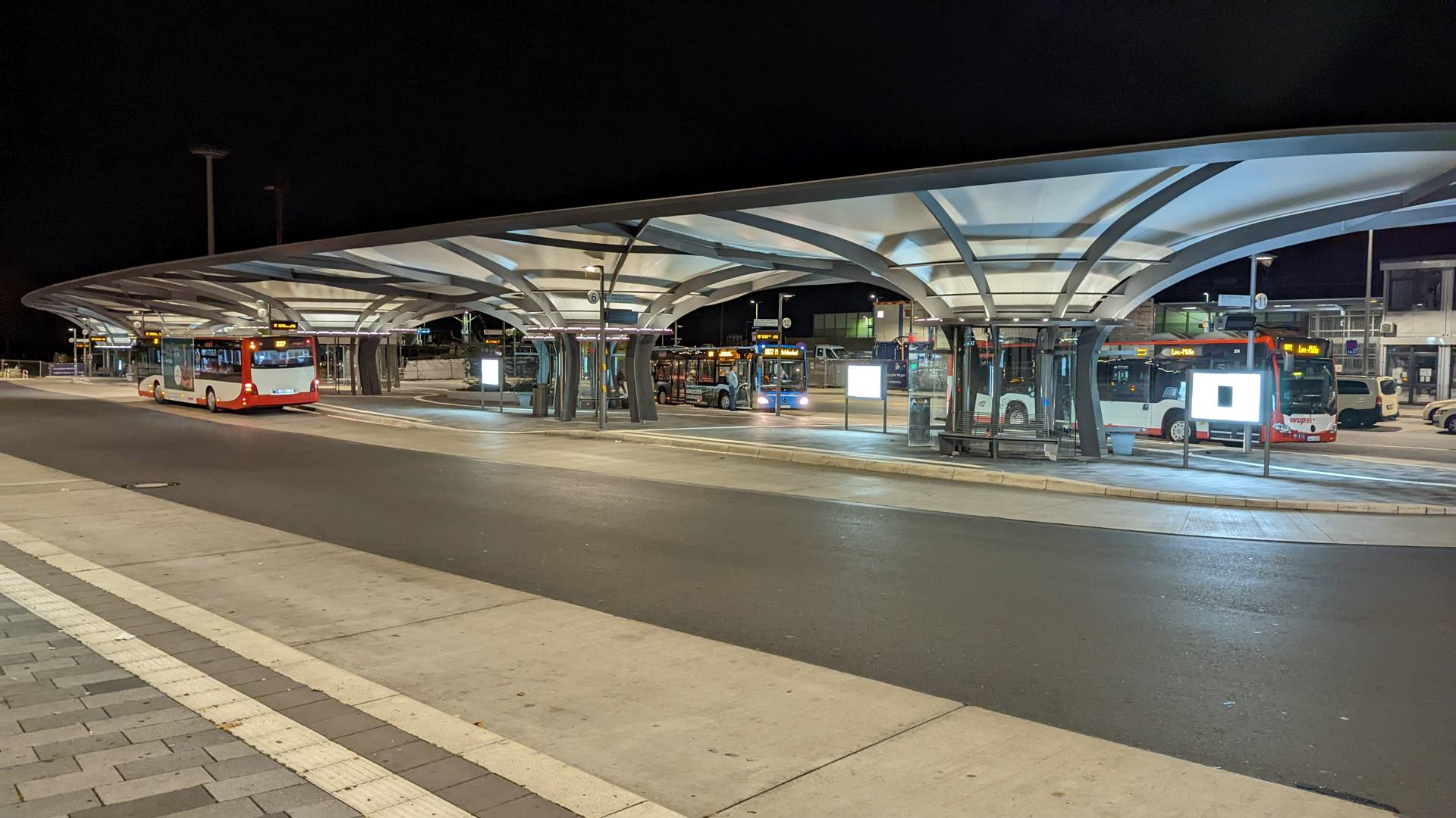 Der neue Busbahnhof in Leverkusen-Mitte. Die Überdachung und Stahlkonstruktion leuchten Stark. An den Bahnsteigen stehen einige Busse, die auf Fahrgäste warten. Im Hintergrund erkennt m an die großen Lichtsäulen, die Leverkusen von weit oben beleuchte