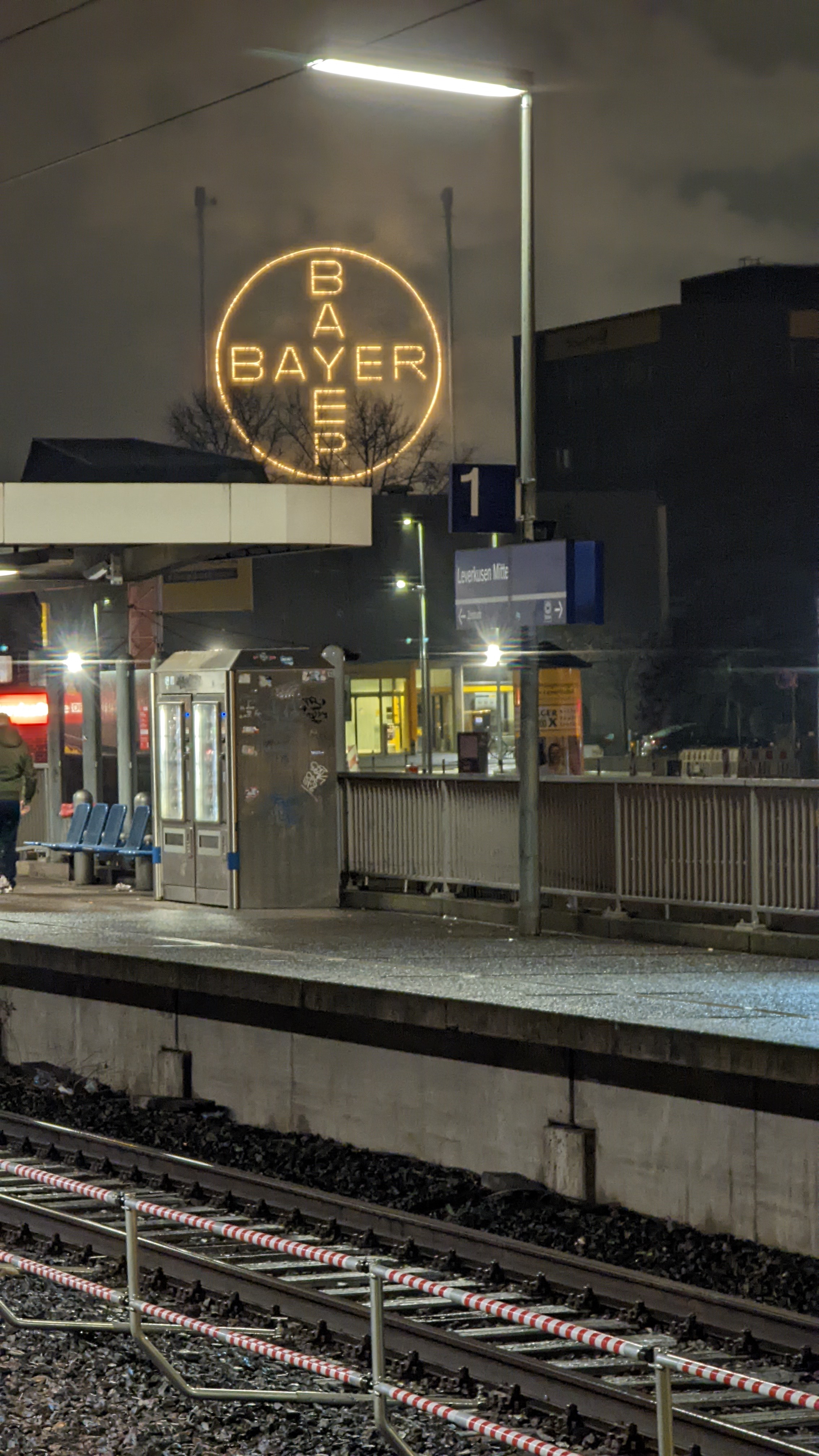 Am unteren Bildrand sind die Schienen und der Bahnsteig von Gleis 1 zu sehen. Der Bahnsteig besteht aus dem Bahnhofsschild "Leverkusen-Mitte", dem Schild "1" für Gleis 1 und zwei Automaten für Snacks und Getränke. Mittig am linken R
